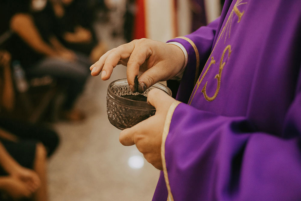A person wearing a purple garment is holding a small, ornate bowl filled with ashes and is about to apply them with their fingers. The background shows people seated, suggesting a religious or ceremonial setting.